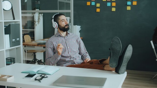 Arab guy is having fun in modern office listening to music pretending to play the guitar wearing wireless headphones relaxing at desk alone. People and lifestyle concept.