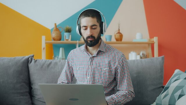 Handsome young Arab guy is working with laptop typing wearing headphones at home. Freelance work, youth culture and apartment activities concept.