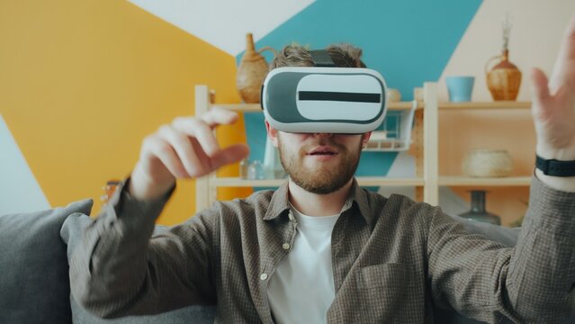 Young man is having fun with vr goggles wearing headset moving arms in apartment in leisure time. Modern technology, people and devices concept.