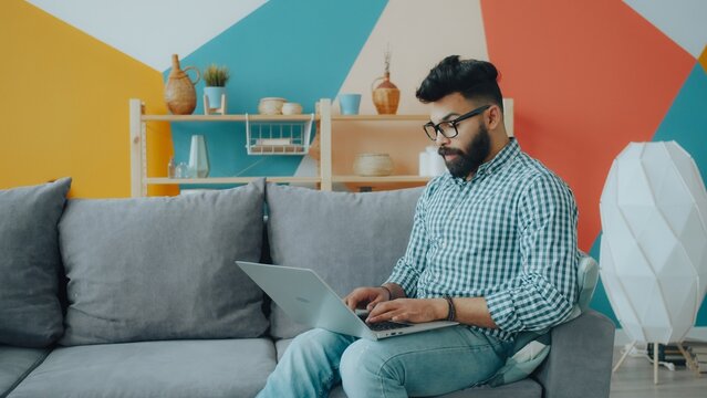 Mixed race guy in glasses and casual clothing is using laptop on sofa in apartment enjoying freelance work. Remote job, people and lifestyle concept.