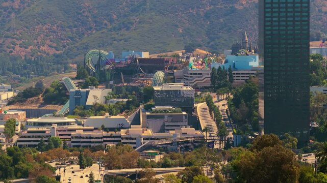 Aerial view of Universal City in Los Angeles California. Theme park roller coasters studio lots palm trees and dark office tower below green Hollywood Hills. Sunny summer day travel landmark.