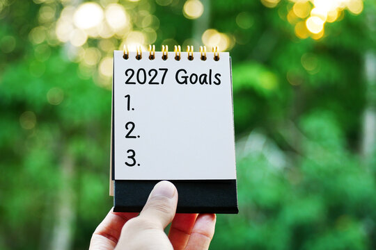 Hand holding spiral notepad with 2027 Goals text and numbered list against green bokeh background