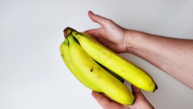 two hands hold a bunch of yellow and green bananas, the fruit rests against a plain white surface, the skin shows small brown spots