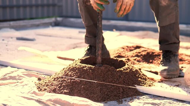 Dusty boots and worker legs stand beside drilling tool as fresh soil spills from mound on covered ground, with bright warm light, blurred corrugated wall, and practical industrial mood.
