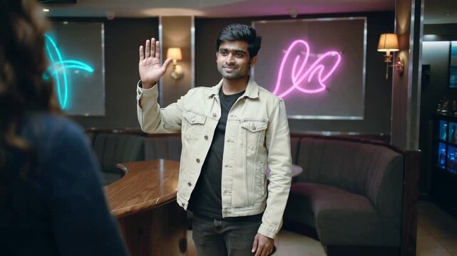 Waving man in beige jacket greeting off-screen guest approaching curved bar, with neon art