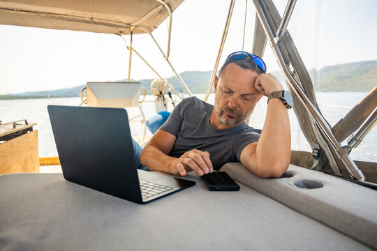 Relaxed man using laptop and smartphone while lounging on a sailing yacht. Digital nomad lifestyle and work life balance at sea.