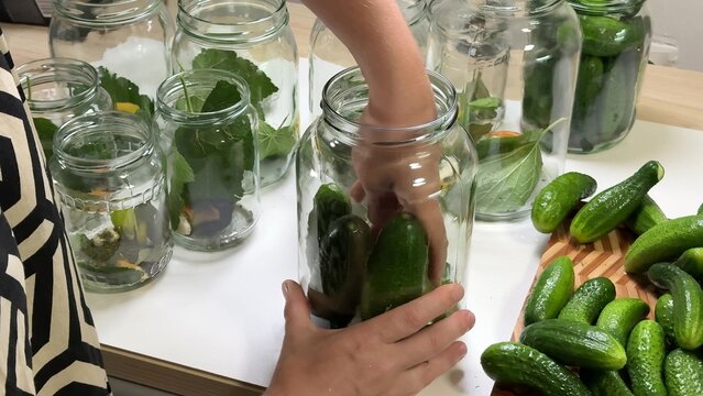 a person handles green cucumbers inside a glass jar, surrounding by other glass jars with leaves and a wooden board with more cucumbers on a white table