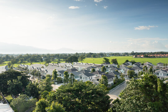 Aerial view of a modern housing estate beside vivid green rice fields in Chiang Mai, Thailand, with tree-lined streets, suburban homes, distant mountains and a bright blue sky in warm afternoon light.