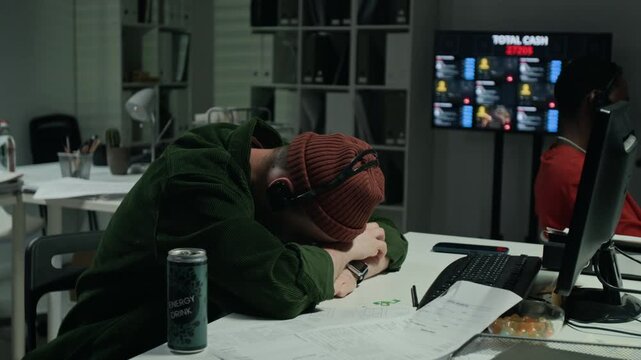 Young Caucasian male hacker wearing headset feeling exhausted at computer desk during night work in scam office. Perfect for cybercrime and scam awareness campaings