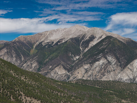 Aerial view of Mount Princeton featuring rugged mountain slopes, dense pine forests, and rocky peaks under a blue sky with wispy clouds in Buena Vista, Colorado, United States.
