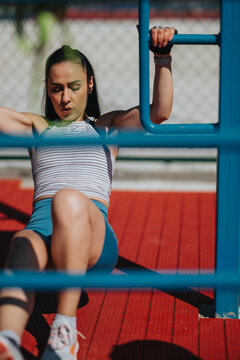 Woman performing a bodyweight core exercise on blue outdoor bars, concentrating on form and effort. The athlete trains outdoors on a red rubber surface using parallel bars for strength work.