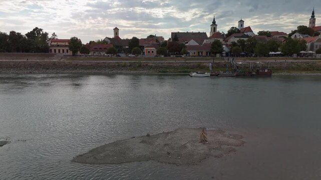 Drone view of a low-water gravel island in the Danube River facing Szentendre's historic waterfront, church towers and moored boats in Hungary during summer.