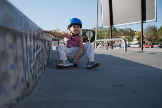 Little Girl in Blue Helmet and Protective Gear Sitting on Penny Board at Skatepark