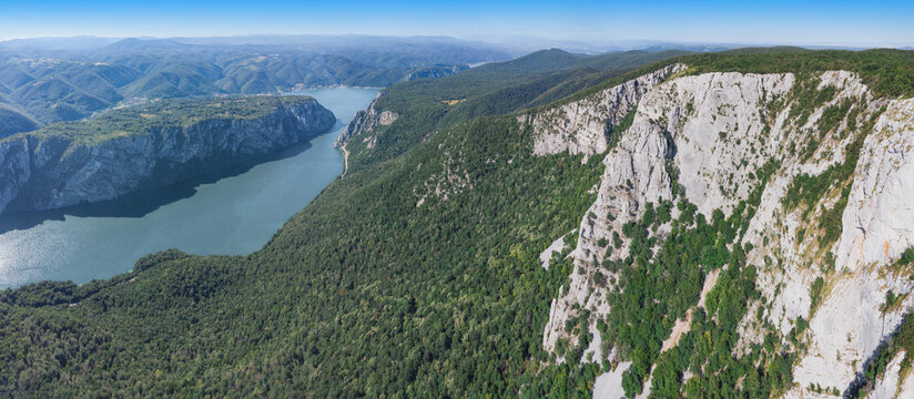 Wide-angle bird's-eye composition centers towering white limestone cliff faces plunging beside a broad river cutting through densely forested gorge ridges. Adventure concept.