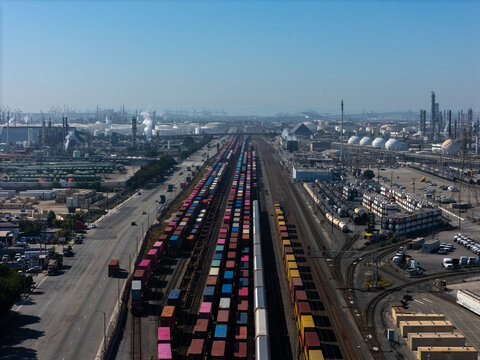 Aerial view of a busy freight railyard with colorful shipping containers on trains, adjacent to an industrial complex with storage tanks in Carson, California, United States.