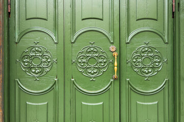 Close up of a carved green wooden door with symmetrical fleur de lis medallion patterns and a vintage brass handle beside an ornate keyhole, decorative entrance detail. © KseniyA