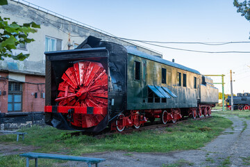 Preserved vintage rotary snowplow locomotive with a large red cutting wheel, displayed on tracks at a railway heritage museum in Dej, Romania, beside old industrial buildings. © Anze