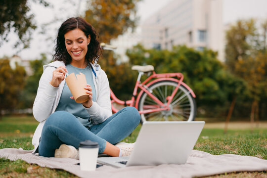 Woman eating takeaway snack while using laptop outdoors