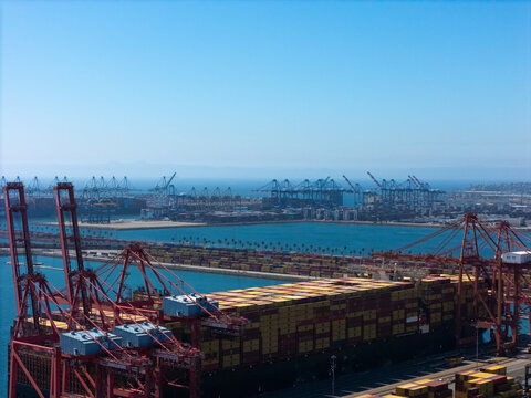 Aerial view of the Port of Long Beach featuring large container ships, red gantry cranes, and stacks of cargo containers under a clear blue sky in Long Beach, California, United States.