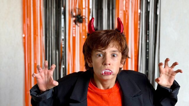 Boy in Halloween costume with red horns and fangs scaring at camera against black‑orange party backdrop (horizontal)