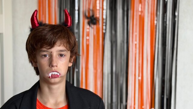  Horizontal shoulder‑up portrait. The subject stands on the left, wearing a black blazer and orange T‑shirt, a red devil horn headband and plastic vampire fangs.