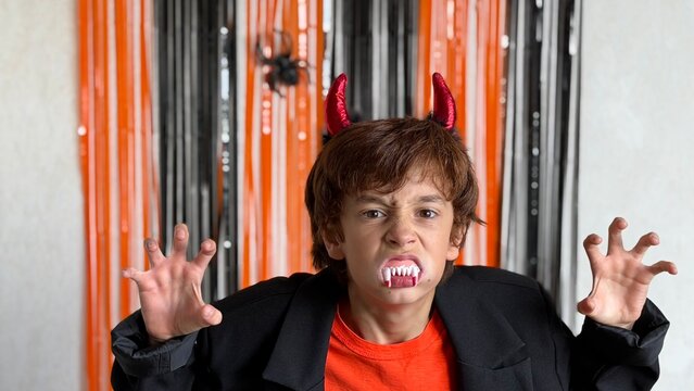 Kid in Halloween costume with red devil horns and vampire fangs snarling at camera against black‑orange streamers