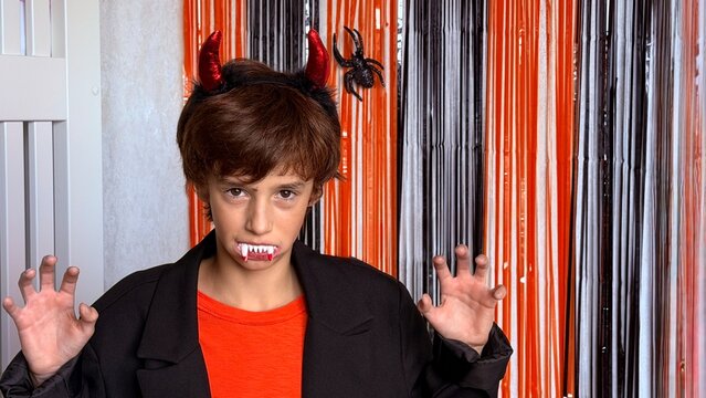 Horizontal medium portrait of a child dressed for Halloween, wearing a shiny red devil horn headband, fake vampire fangs, an orange T‑shirt and a black blazer