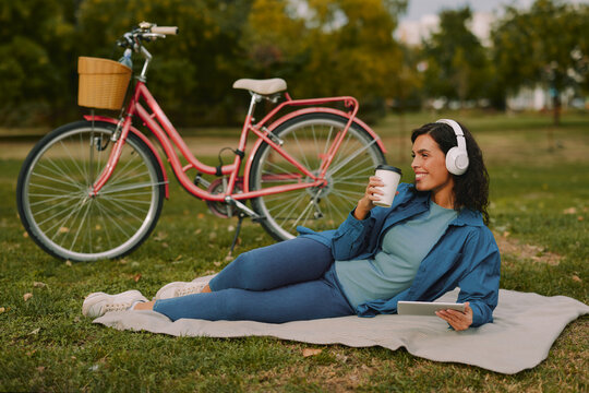 Woman relaxing with headphones, coffee and tablet in park