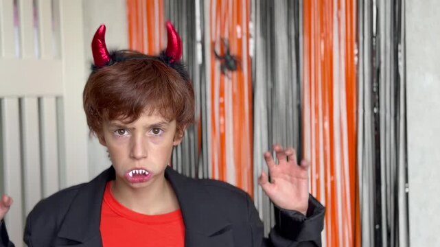  The kid looks into the lens, raises claw hands and makes a scary face. Background features vertical black and orange streamers with a decorative spider
