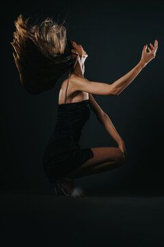 A young woman in a black dress flips her hair while kneeling in a dark studio. The dramatic movement and lighting emphasize motion and an elegant, expressive pose.