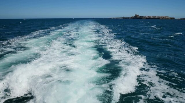 Fast boat movement creating sea foam, white water, and swirling waves in the Mediterranean Sea. Scenic view of Tabarca Island in the background under bright natural sunlight in Alicante, Spain.