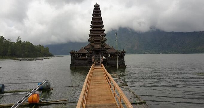 A drone flies low along a wooden bridge toward the eleven-tiered meru of Pura Segara Ulun Danu Batur under a cloudy sky on Lake Batur, Aerial