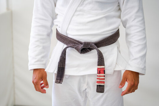Close up of a worn Brazilian jiu jitsu brown belt tied over a white gi, with rank stripes on one end and the hands of the practitioner at the sides