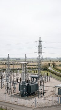 High voltage power transformer station and electrical transmission tower standing in rural countryside landscape under overcast sky background