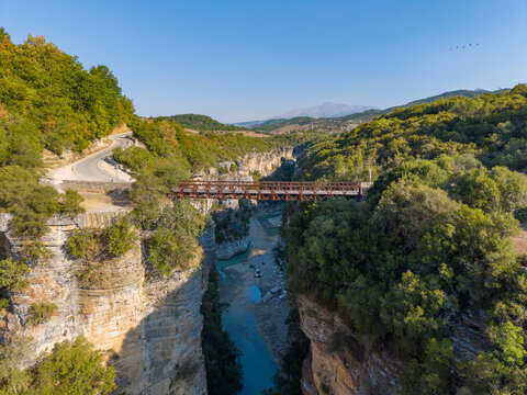 Aerial view of Osumi Canyon featuring a metal bridge spanning the deep gorge with winding roads and lush green vegetation under a clear blue sky in Dhores, Berat County, Albania.