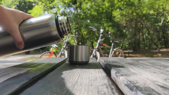 Pouring hot tea from thermos into metal cup on wooden picnic table in park. Traveler serving hot beverage from steel flask during outdoor recreation, with parked bicycles visible on background.