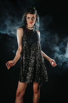 Young woman in a black sequin dress with decorative face gems poses against a smoky dark background. She stands confidently in festival makeup and glitter for a nighttime party portrait.