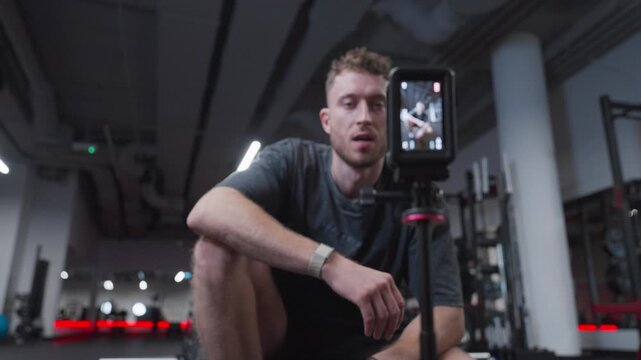 An athletic man checks an action camera and then moves into position beside a barbell for a gym video shoot.