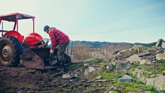 Person Loads Large Rocks Into The Front Loader Bucket Of A Red Farm Tractor On A Rural Hillside. Static Shot