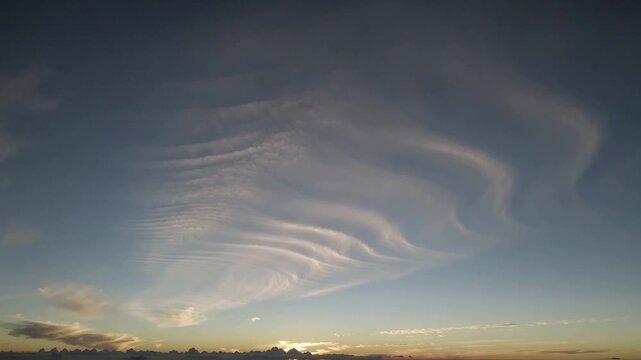 Shifting rippled cloud bands spreading right as sun emerging behind horizon bank, reflecting gold