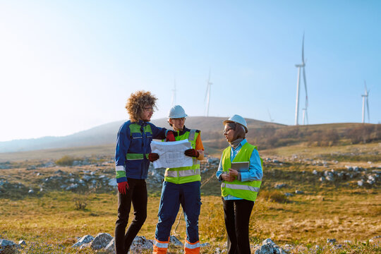 Engineers Reviewing Wind Farm Plans On Site With Safety Vests And Hard Hats For Renewable Energy Project
