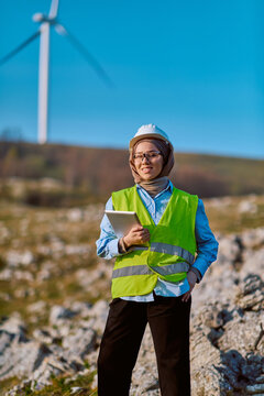 Female Engineer in Hijab Wearing Safety Vest and Hard Hat Inspecting Wind Turbine With Tablet on Wind Farm