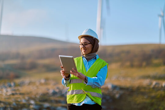 Female Engineer Wearing Hard Hat And High-Visibility Vest Inspecting Wind Turbine Site With Tablet