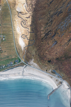Aerial view of the rugged coastline of Senja island, featuring a winding stream, steep mountain slopes, a sandy beach, and a coastal road in Senja, Troms, Norway.
