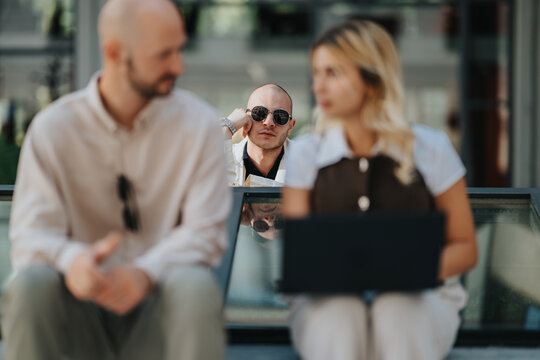 Man in sunglasses leans on a railing while looking toward two people sitting nearby. A blurred couple uses a laptop on outdoor steps with an onlooker in the background.