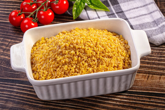 Cooked bulgur wheat in white ceramic baking dish with cherry tomatoes and basil on wooden table