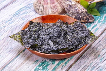 Weathered blue and white wooden surface with dark nori seaweed sheets in terracotta bowl, seashell and basil leaf nearby © Andrei Starostin