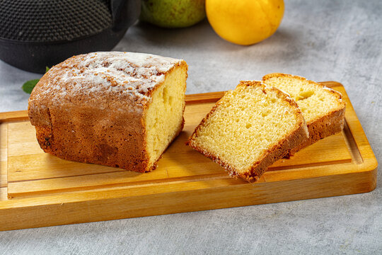 Wooden cutting board holding sliced pound cake dusted with powdered sugar, golden crumb visible, yellow fruit and dark kitchenware in background