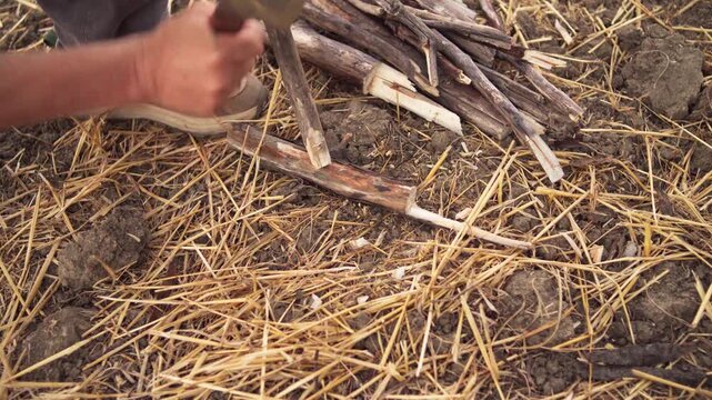 A camper sharpens a wooden stake with a heavy cleaver knife on straw ground to hang a pot over the fire, close-up shot.