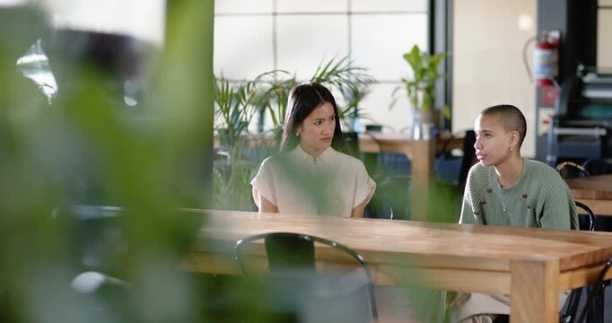 Asian, African American women sitting at wooden table, right speaker initiating talk left reacting